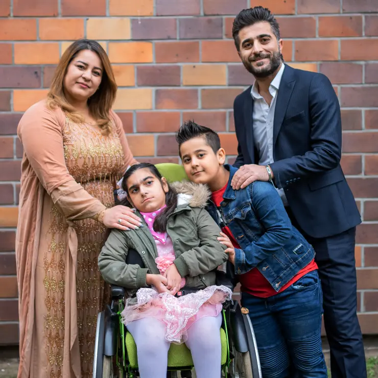 Eine Familie von 4 Personen steht vor einer Ziegelmauer. Links die Mutter, rechts der Vater. In der Mitte sitzt die Tochter im rosa Tüllkleid im Rollstuhl. Ihr Bruder lehnt sich zu ihr. Die Eltern haben ihre Hände auf den Schultern der Kinder.