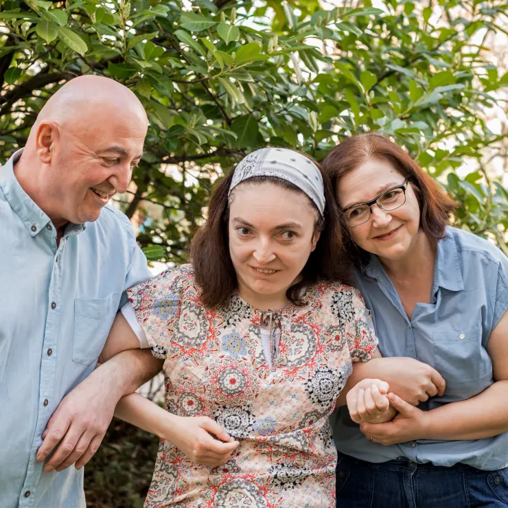 Eine Familie von 3 Personen steht vor einem Baum mit grünen Blättern. Vater und Mutter haben ihre Tochter eingehakt. Die Tochter steht in der Mitte. Sie trägt in ihrem mittellangen Haar ein Tuch. Ihre helle Bluse ist geblümt. Der Vater links schaut lächelnd zu seiner Tochter. Die Mutter rechts legt ihren Kopf an den Kopf ihrer Tochter.