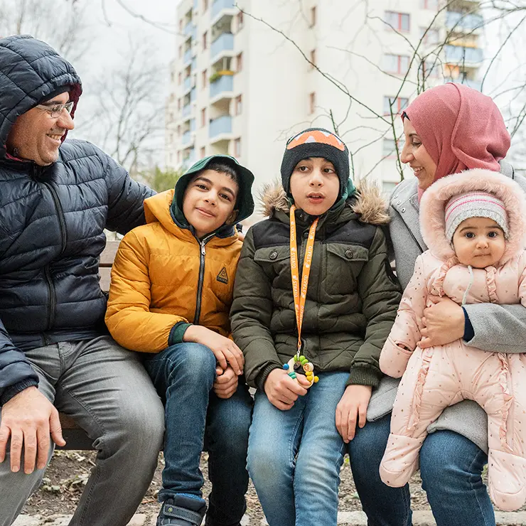 Eine Familie von 5 Personen sitzt auf einer Bank. Hinter ihr ein hohes Wohnhaus. Es ist Winter. Alle haben dicke Jacken an und eine Kopfbedeckung auf. Links sitzt der Vater, er schaut lächelnd auf seine beiden Söhne. Rechts sitzt die Mutter. Sie hat ein Baby auf dem Arm. Zwischen den Eltern sitzen zwei Jungen. Einer hat an einem Band um den Hals eine Kinderrassel.