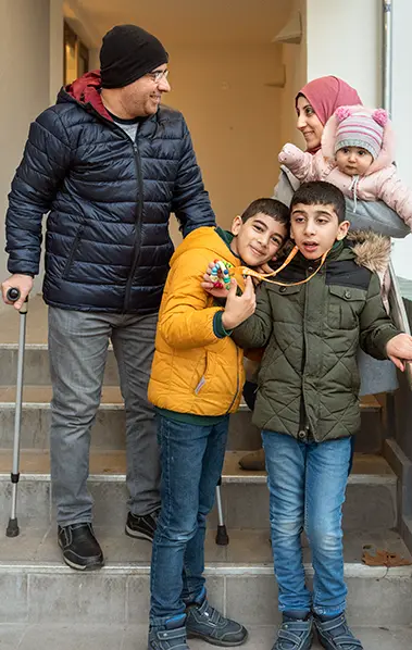 Die Familie steht auf der Treppe. Links der Vater, er stützt sich auf Krücken. Rechts die Mutter, sie hält das Baby im Arm. Vorne stehen die beiden Jungen. Einer hält die Rassel hoch. Sein Bruder lehnt den Kopf an seinen.