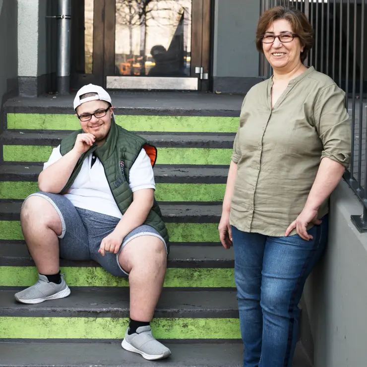 Mutter und erwachsener Sohn vor einem Hauseingang mit Stufen. Die Mutter steht vorne rechts. Sie lehnt lächelnd an einem Geländer. Sie trägt Jeans und eine olivgrüne Bluse. Der Sohn sitzt auf den Treppenstufen neben ihr. Er trägt eine kurze Hose, ein weißes Poloshirt und eine grüne Weste. Er hat seinen Ellenbogen auf ein Bein gestützt. Mit seiner Hand am Kinn lächelt er in die Kamera.