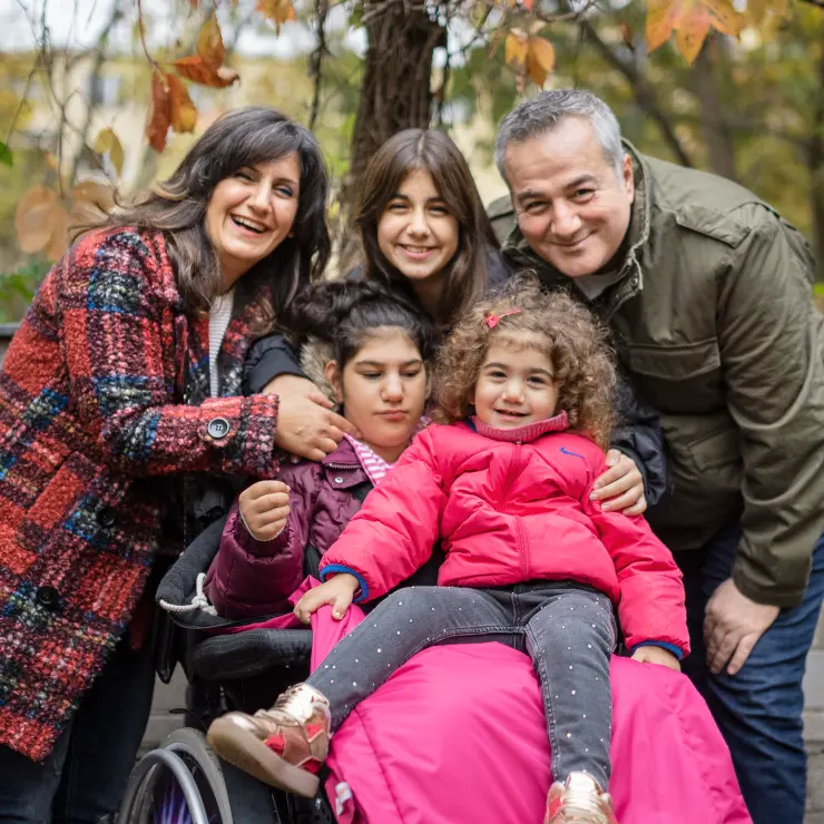 Die 5-köpfige Familie steht vor einer Kastanie im Herbst. Der Vater ist links, die Mutter rechts, dazwischen 3 Töchter. Die älteste Tochter sitzt unter einer pinkfarbenen Decke im Rollstuhl. Auf ihrem Schoß sitzt ihre kleine Schwester in einer Jacke in Pink. Hinter dem Rollstuhl steht die dritte Tochter. Sie und ihre Eltern beugen sich zu den beiden herunter. Sie lächeln in die Kamera.