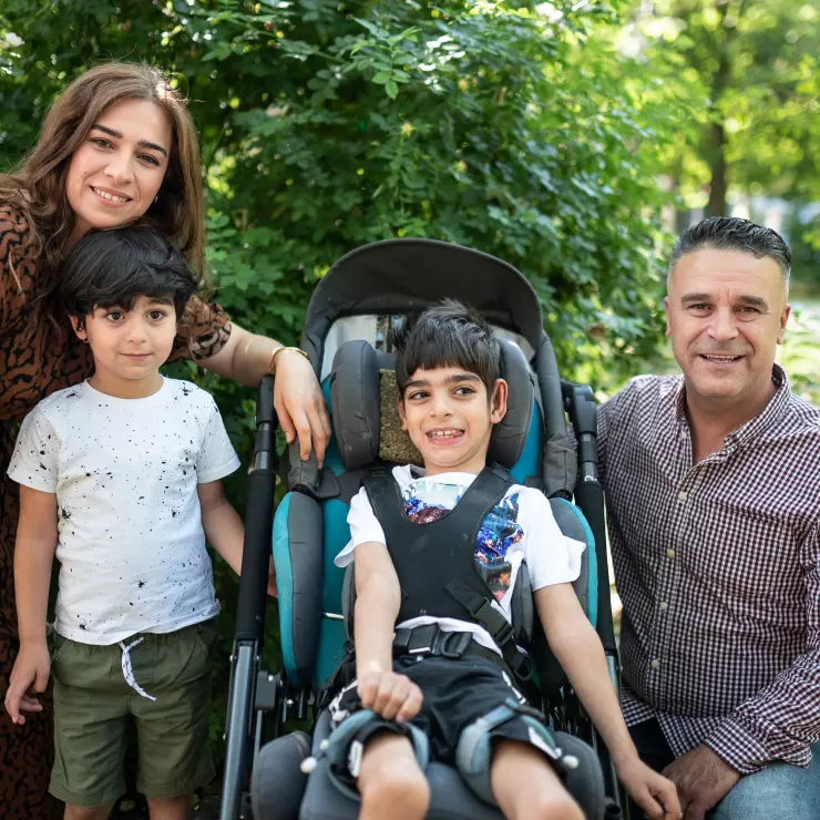 Eine 4-köpfige Familie steht im Park vor grünen Blättern. In der Mitte sitzt ein kleiner schmaler Junge im Rollstuhl. Er grinst und freut sich. Sein Vater hat sich links neben ihn gehockt. Rechts von ihm steht sein kleiner Bruders. Dahinter die Mutter. Sie beugt sich herab. Ihr Kinn ruht auf dem Kopf des kleines Bruders.