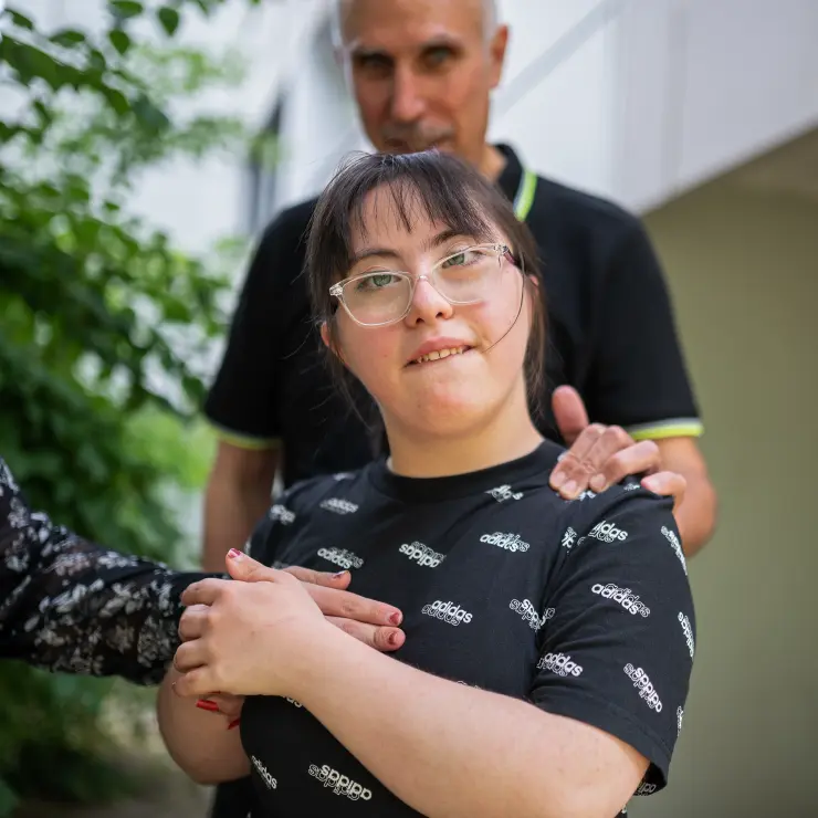 Im Mittelpunkt steht eine junge Frau. Sie trägt eine helle Brille und ein schwarzes T-Shirt. Darauf steht überall der Schriftzug von adidas. Im Hintergrund ist unscharf ihr Vater zu sehen. Er hat eine Hand auf die Schulter seiner Tochter gelegt. Die Hand einer Frau kommt von vorne. Diese Hand hat die Tochter ergriffen und drückt sie sich auf die Brust. Die Frau und die Tochter tragen roten Nagellack.