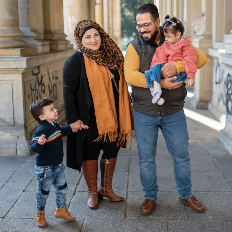 Eine 4-köpfige Familie steht in einem Säulengang. Der Vater ist links im Bild. Er hat seine kleine Tochter auf dem Arm. Neben ihm steht die Mutter. Sie trägt ein braun-schwarzes Kopftuch und braune Stiefel. An ihrer rechten Hand hält sie ihren kleinen Sohn. Der schaut hoch zum Vater. Die beiden lächeln sich an.