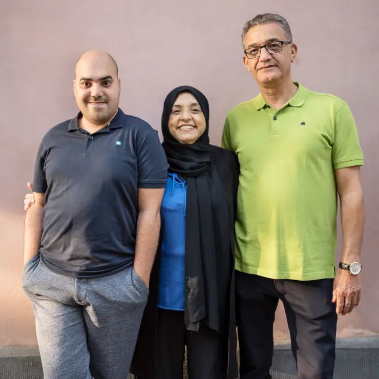 Eine Familie von 3 Personen steht vor einer hellen Hauswand. Die Mutter ist die kleinste Person. Sie trägt ein schwarzes Kopftuch und steht in der Mitte. Rechts von ihr ist der Vater, links der Sohn. Die Mutter hat beide mit ihren Armen umfasst. Der Sohn steht lässig mit Händen in den Taschen.