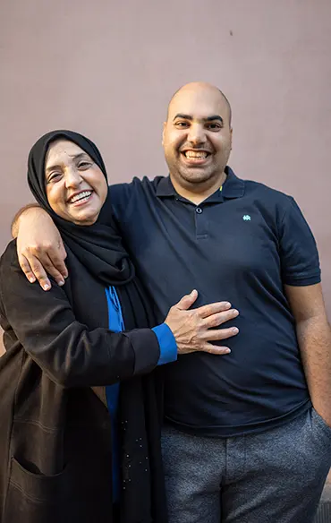 Mutter und Sohn stehen lachend vor der Hauswand. Sie wirken glücklich und entspannt. Er hat seinen rechten Arm um ihre Schulter gelegt. Die andere Hand steckt in seiner Hosentasche. Die Mutter hat ihre rechte Hand auf seine Brust gelegt.