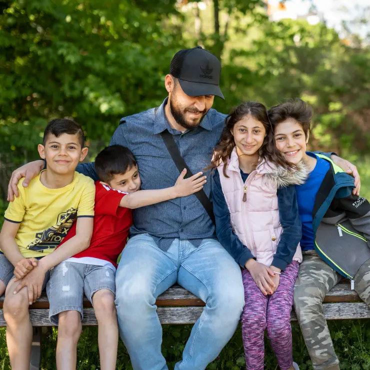 Eine 5-köpfige Familie sitzt auf einer Holzbank. Im Hintergrund sind verschwommen Bäume und grüne Blätter zu sehen. Der Vater sitzt in der Mitte. Er hat seinen beiden Arme um seine Kinder gelegt. Links sitzen die kleinsten. Es sind zwei Jungen. Der kleine Junge direkt neben ihm umarmt seinen Vater. Links sitzen die älteren beiden Kinder. Ein Mädchen und ein Junge. Der Junge lehnt sich an das Mädchen.