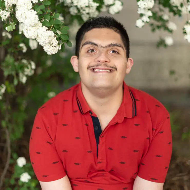 Ein junger Mann strahlt in die Kamera. Sein Oberkörper bis zu den Armen ist zu sehen. Er sitzt unter einem Baum mit weißen Blüten. Er trägt ein rotes Poloshirt.