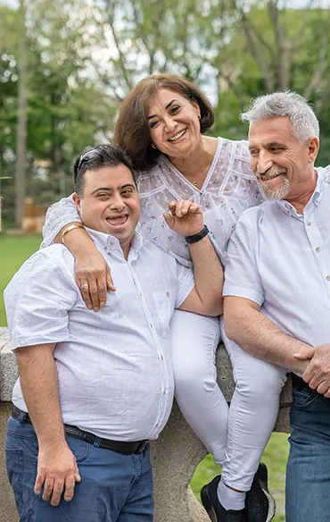 Die Mutter sitzt auf einer Steinmauer. Sohn und Mann stehen links und rechts von ihr. Sie hat ihre Arme über die Schultern der beiden gelegt. Die beiden lehnen sich an die Mauer und zu ihr. Alle lächeln.