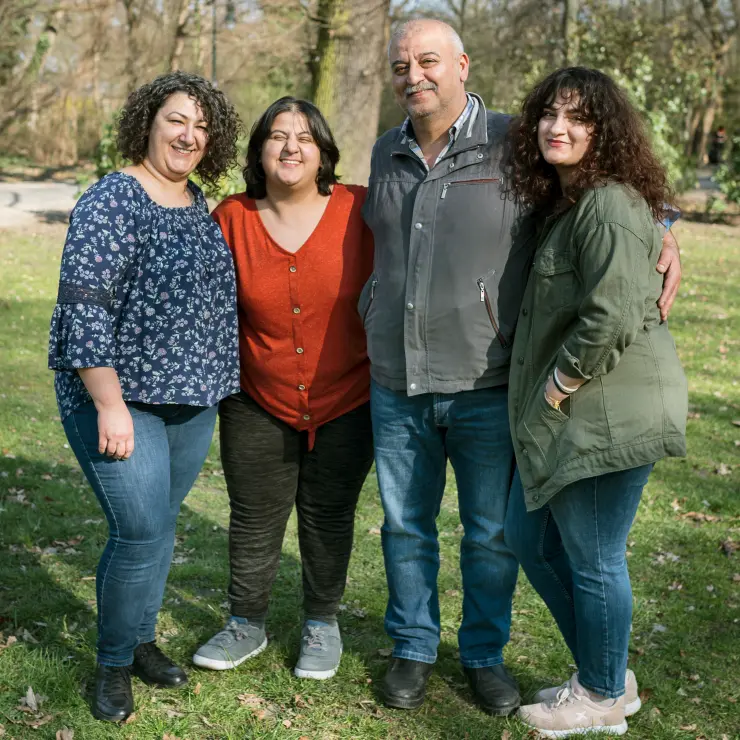 Eine 4-köpfige Familie steht auf der Wiese. Es sind Vater, Mutter und zwei erwachsene Töchter. Die Mutter steht ganz links. Dann eine Tochter mit rotem Oberteil. Daneben der Vater und ganz rechts die zweite Tochter. Sie stehen eng nebeneinander und haben die Arme umeinander gelegt. Die Sonne scheint ihnen ins Gesicht. Deshalb kneifen sie etwas die Augen zusammen.